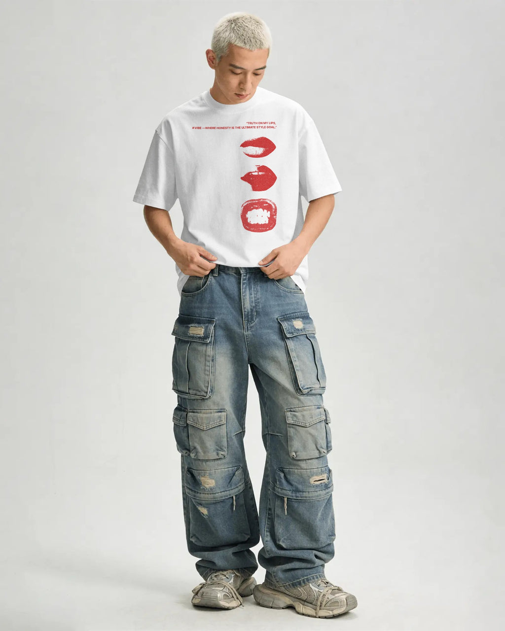 A model with platinum blonde hair stands against a light gray studio background wearing a white graphic t-shirt with red lips, blue denim cargo pants, and gray sneakers.