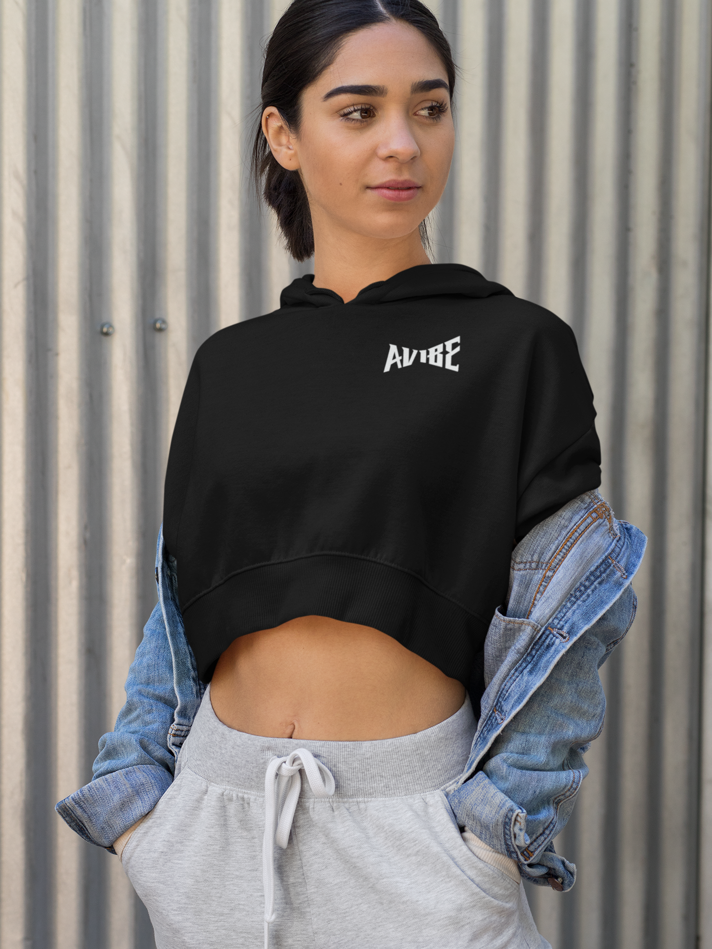 Woman wearing a black hoodie with 'AVIE' logo, gray pants, and denim jacket against a corrugated metal wall.