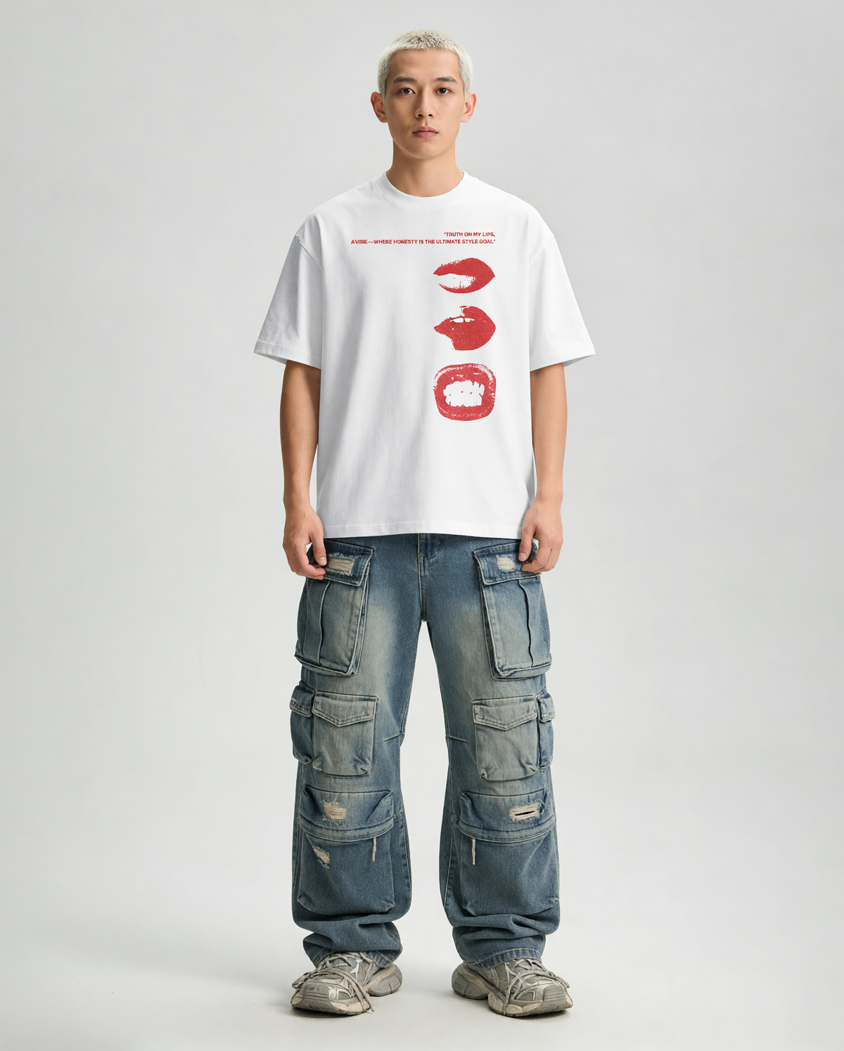A model stands against a light gray studio background wearing a white graphic T-shirt with red lip and hand illustrations, blue cargo pants, and gray sneakers.
