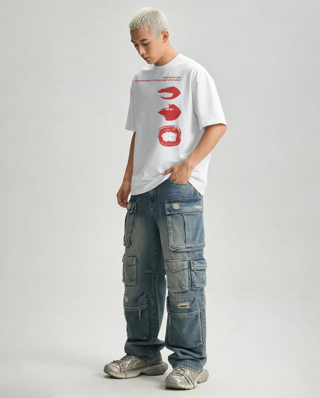 A model with short blonde hair is wearing a white graphic t-shirt with red lips, blue denim cargo pants, and gray sneakers, standing against a light gray studio background.