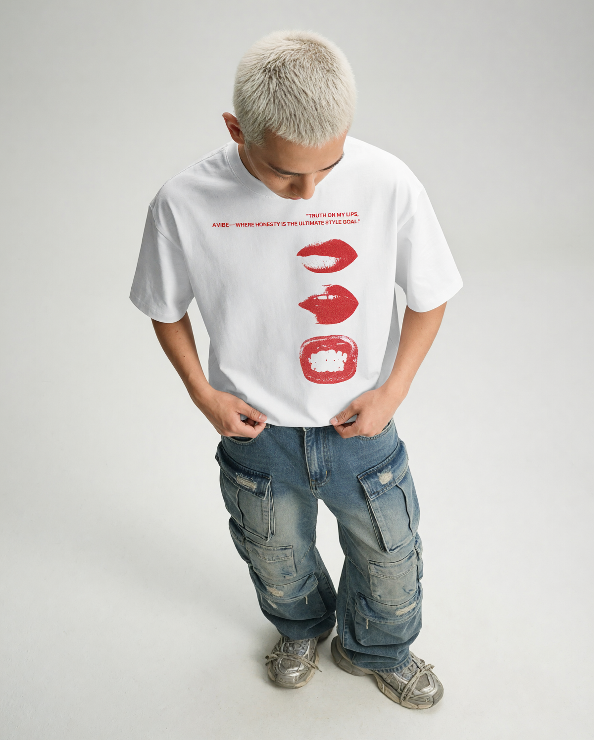 A child model wearing a white graphic t-shirt with red lips and text, paired with blue cargo jeans and gray sneakers, standing against a light gray studio background.