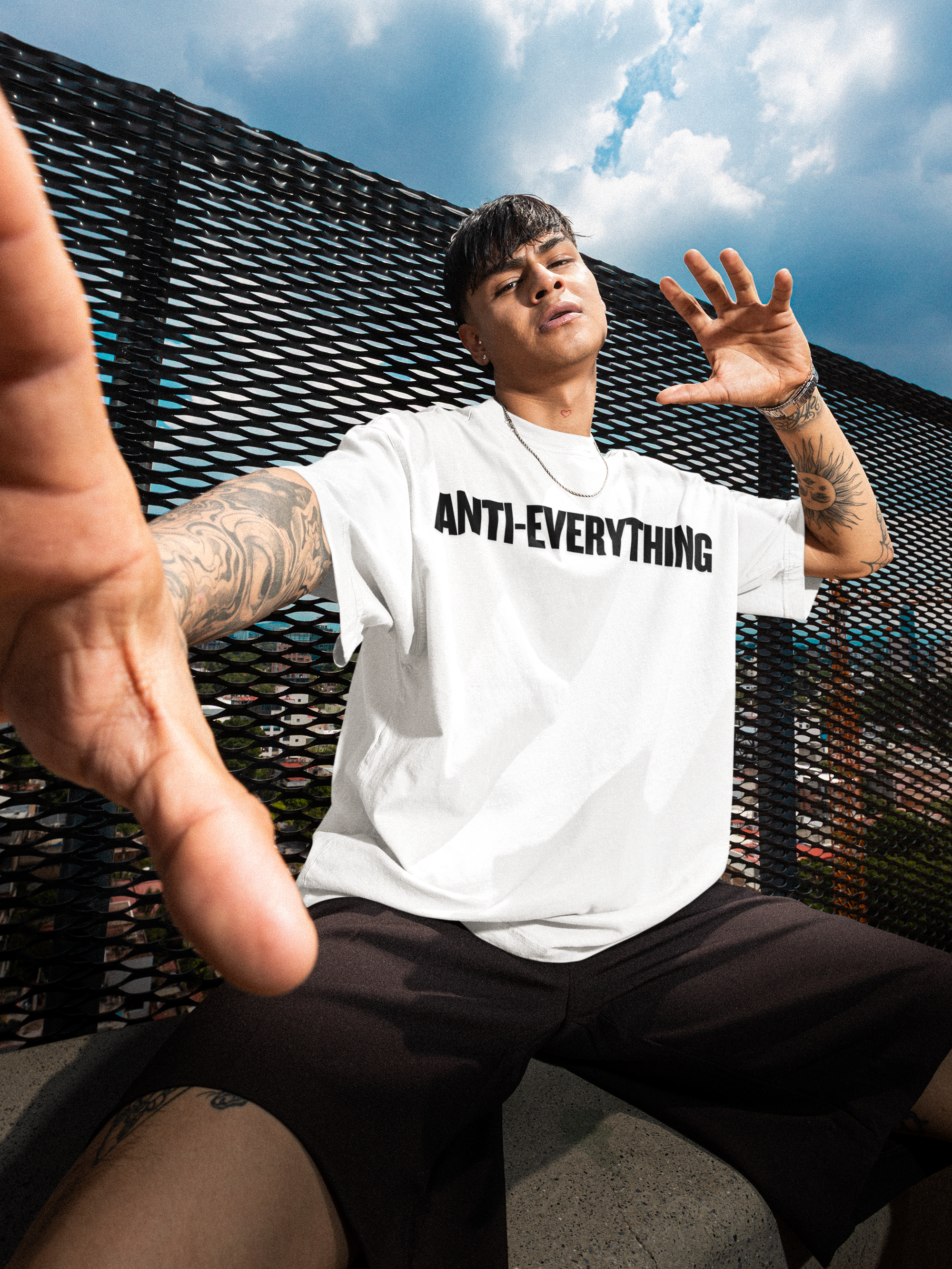 Person wearing a white t-shirt with 'ANTI-EVERYTHING' text, sitting outdoors with a textured wall and blue sky in the background.